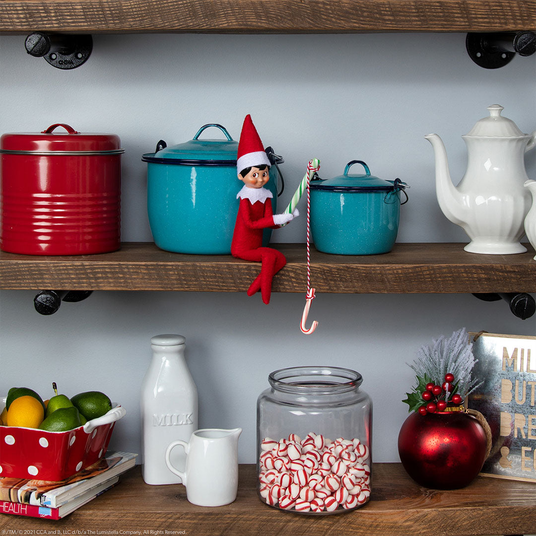 Elf on the Shelf elfje zit op een plank tussen keukengerei en houdt een zuurstok vast.