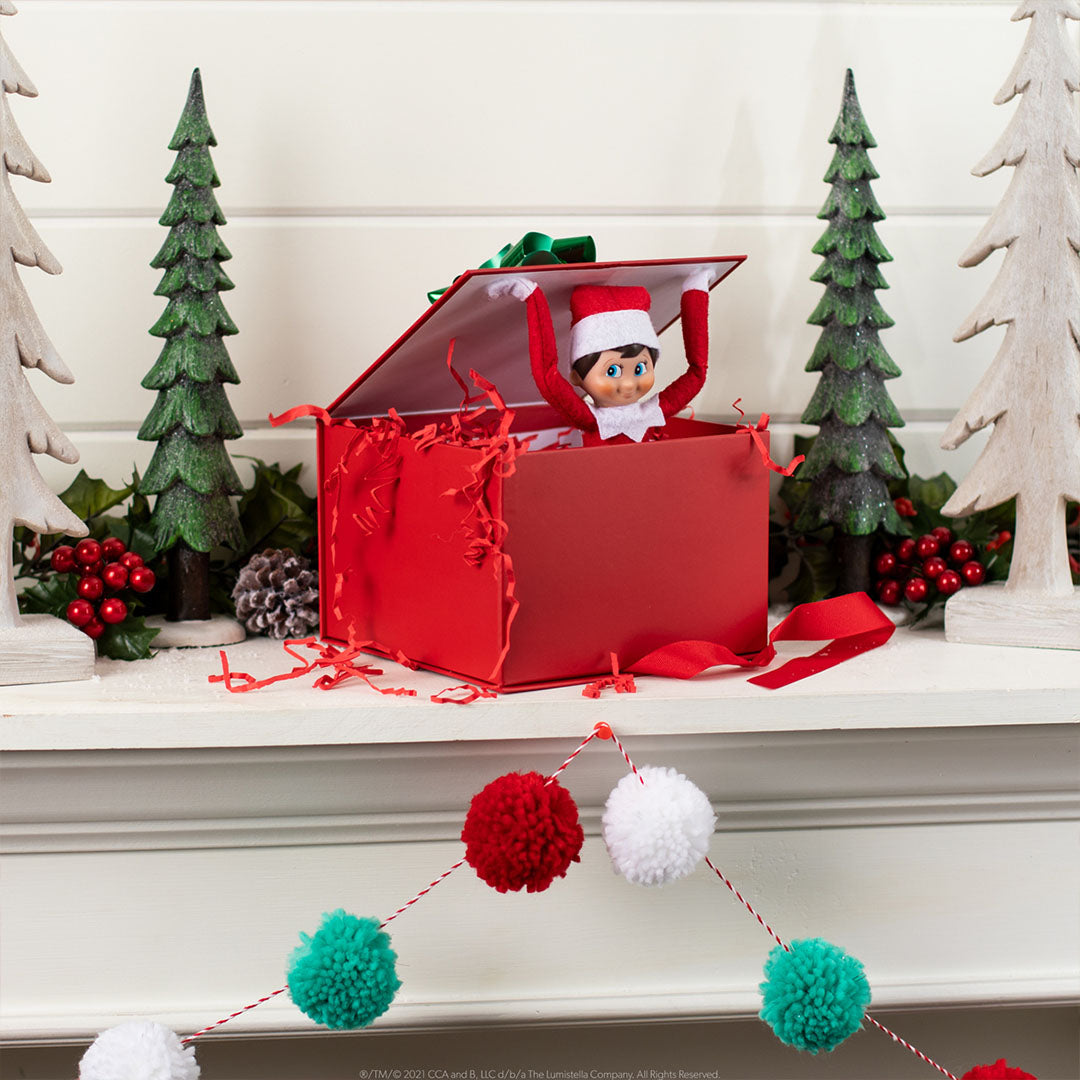 Elf on the Shelf elfje springt uit een rode cadeaubox op een kerstversierde schoorsteenmantel.