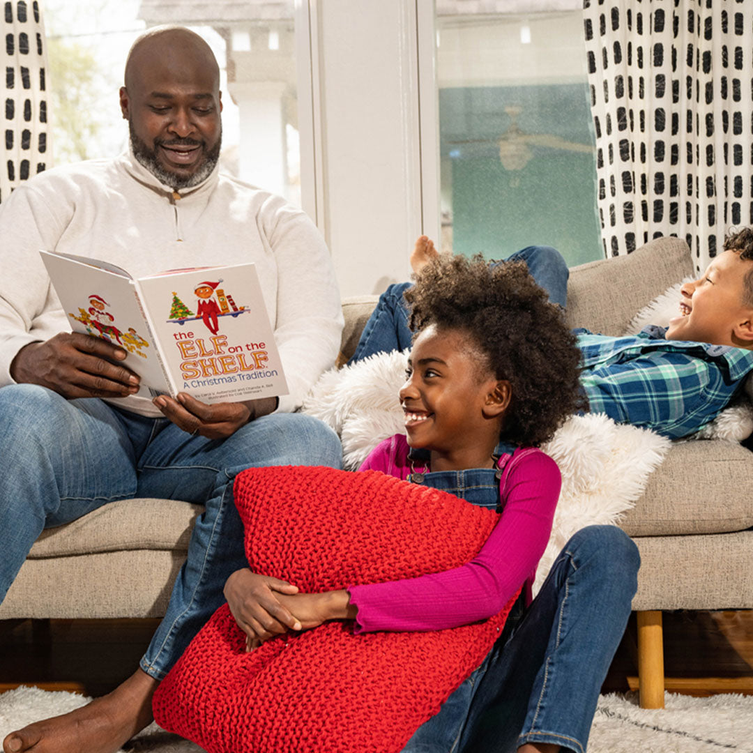 Een man leest het Elf on the Shelf kerstboek voor aan twee lachende kinderen op de bank.