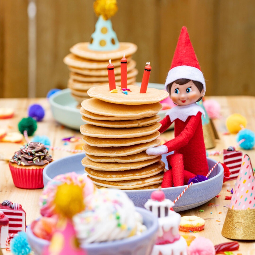 The Elf on the Shelf elfje zit naast een stapel pannenkoeken met kaarsjes op een feestelijk gedekte tafel.