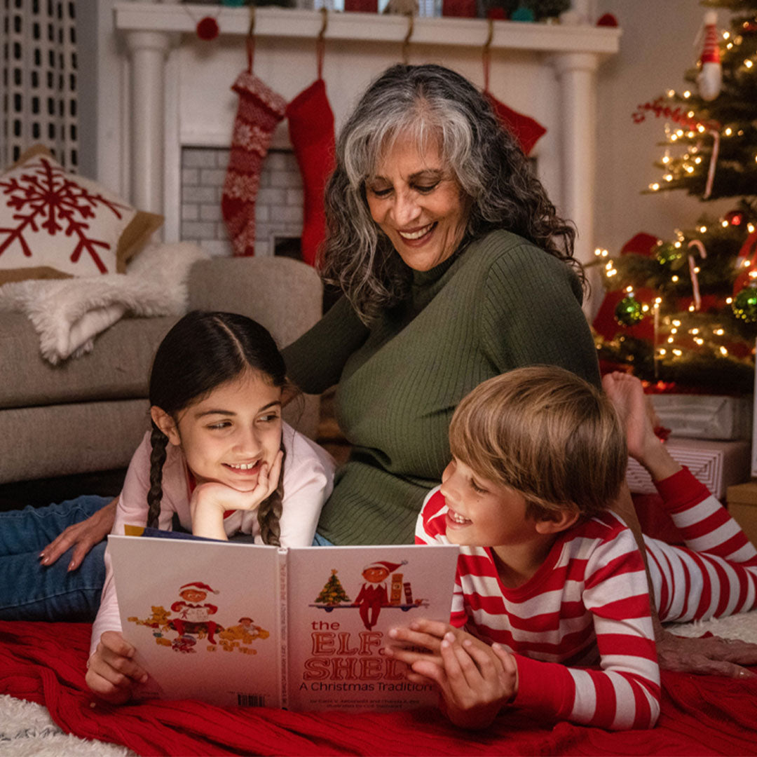 Een vrouw en twee kinderen lezen samen het The Elf on the Shelf kerstboek voor een verlichte kerstboom.