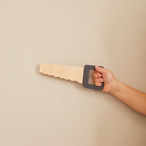 Een kinderhand houdt een houten speelgoedzaag vast tegen een lichte muur.