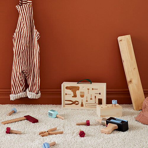 Kids Concept houten gereedschapskoffer en losse gereedschapsstukken liggen op een wit tapijt voor een oranje muur.