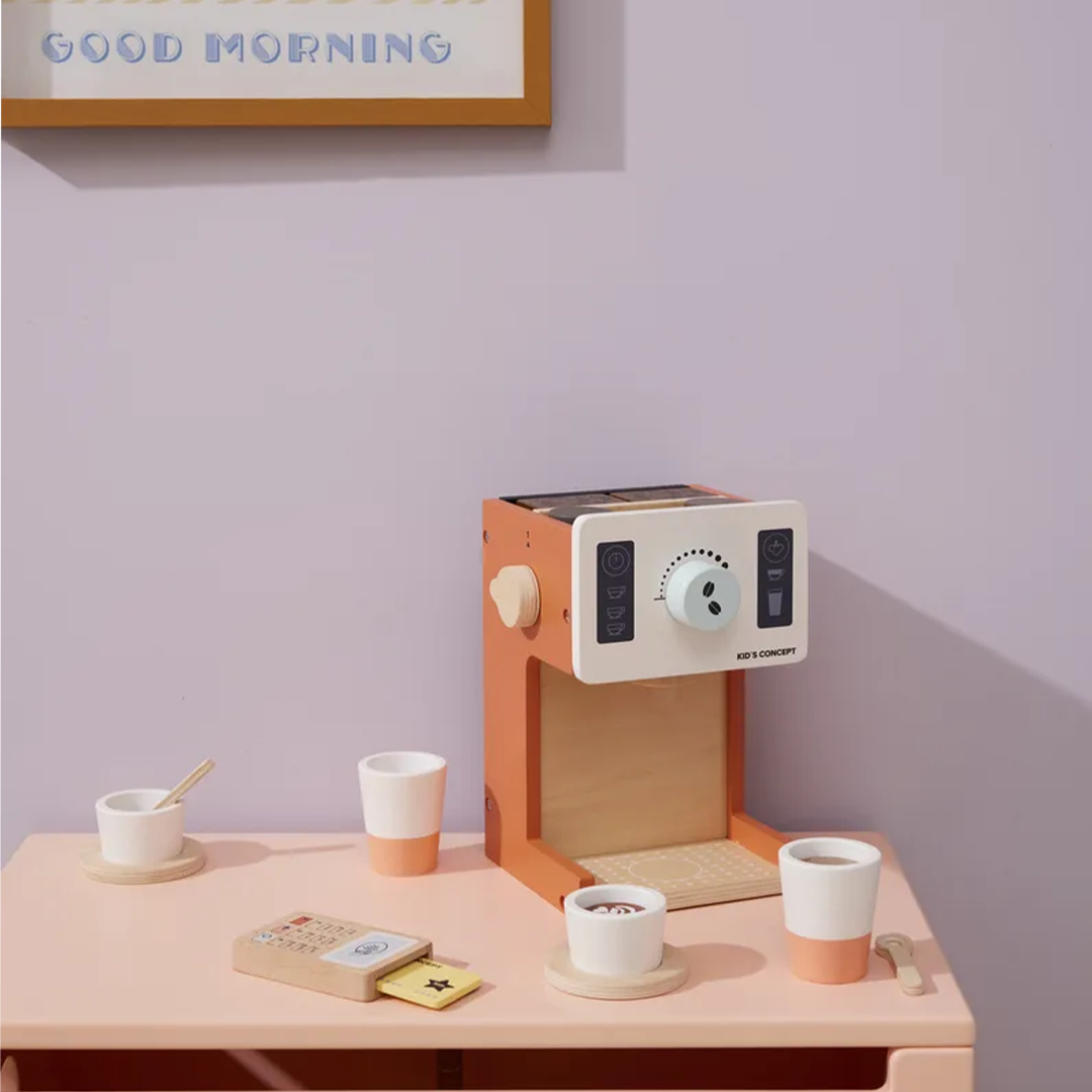 Een Kids Concept houten koffiemachine speelset met kopjes en een betaalautomaat op een roze tafel, met een paarse muur en een lijst met 'Good Morning' op de achtergrond.
