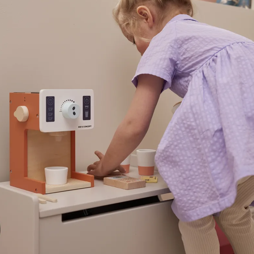 Een kind in een paarse jurk speelt met de Kids Concept houten koffiemachine speelset op een witte bank.