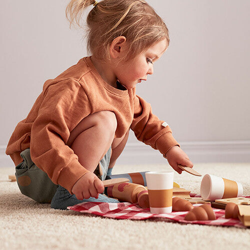 Een kind speelt op de grond met de houten picknick speelset, inclusief speelgoedeten en een rood geruite mat.