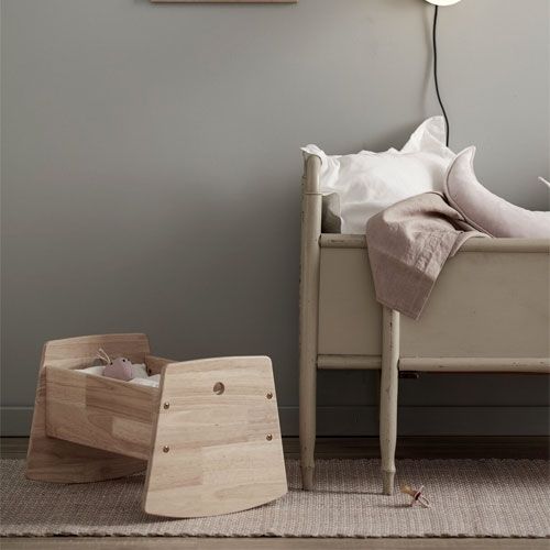 Kids Concept houten poppenwieg met een knuffeltje erin, staand op een vloerkleed naast een groot bed.