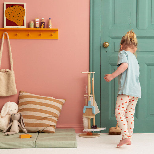 Een kind staat naast de Kids Concept houten schoonmaakset Kid's Hub in een kamer met een roze muur en een groene deur.