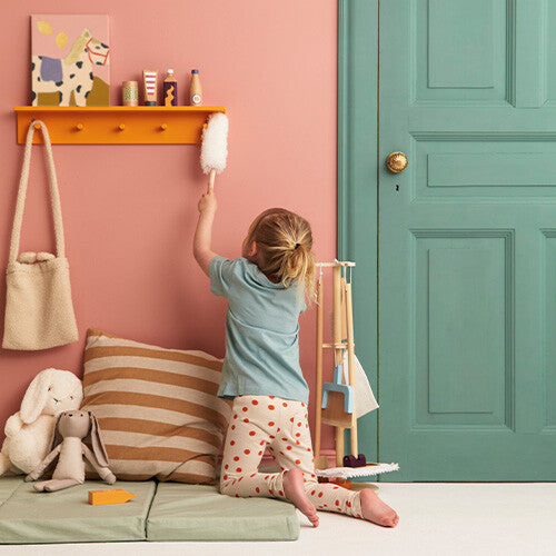 Een kind knielt en veegt met de duster van de Kids Concept schoonmaakset tegen een roze muur.