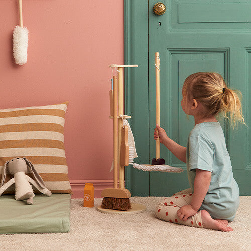 Een kind knielt en houdt de houten bezem van de Kids Concept schoonmaakset vast naast de standaard.