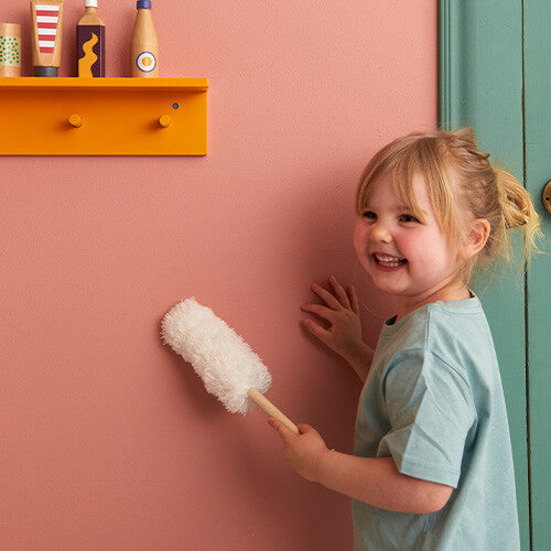 Een lachend kind houdt de duster van de Kids Concept schoonmaakset vast tegen een roze muur.