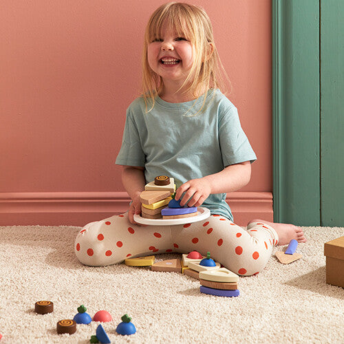Een lachend kind zit op de grond en houdt een Kids Concept houten speelgoedtaart vast, met losse stukken eromheen.