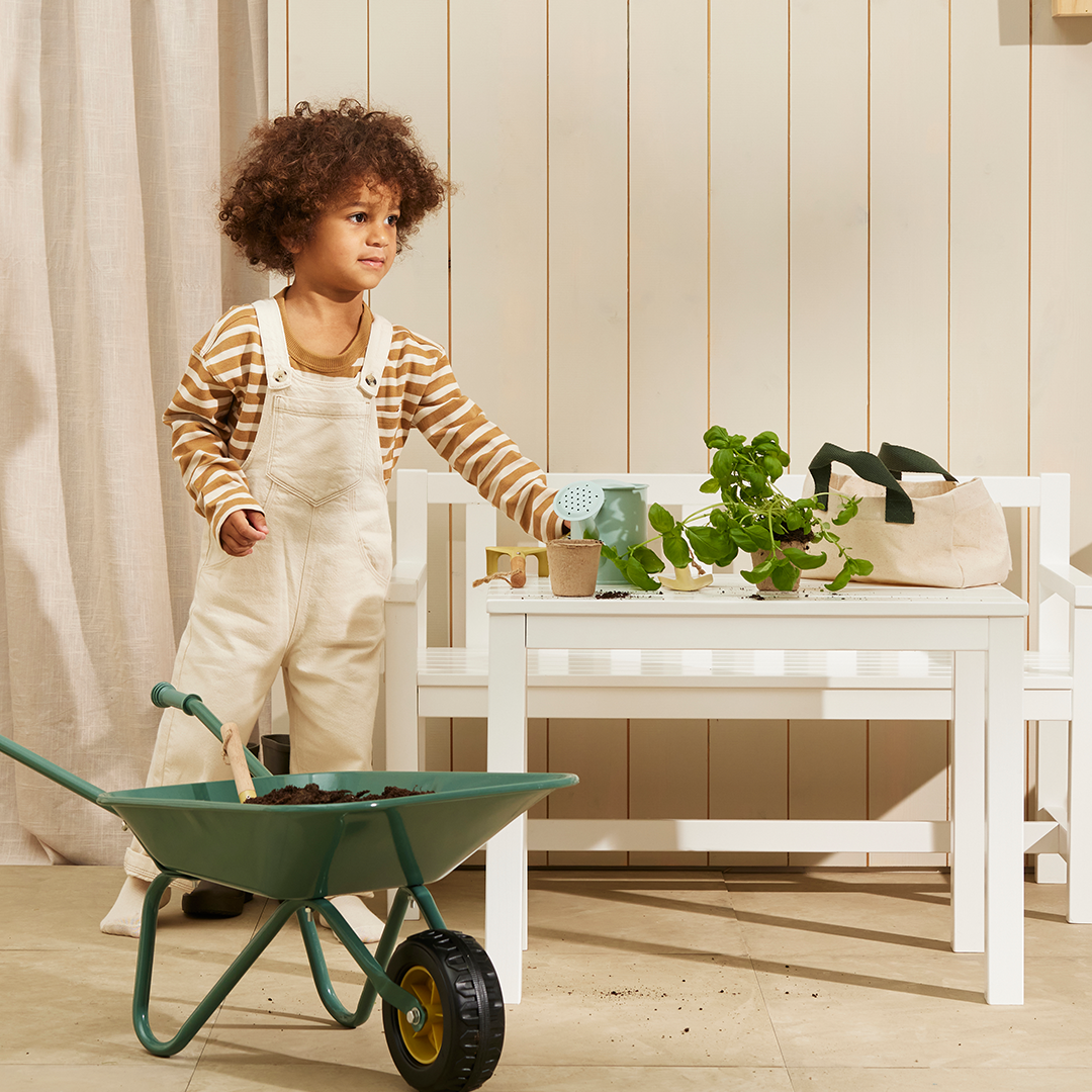 Een jongen met krullen giet water op een plantje met de Kids Concept tuingereedschapsset, naast een kruiwagen en een witte bank.