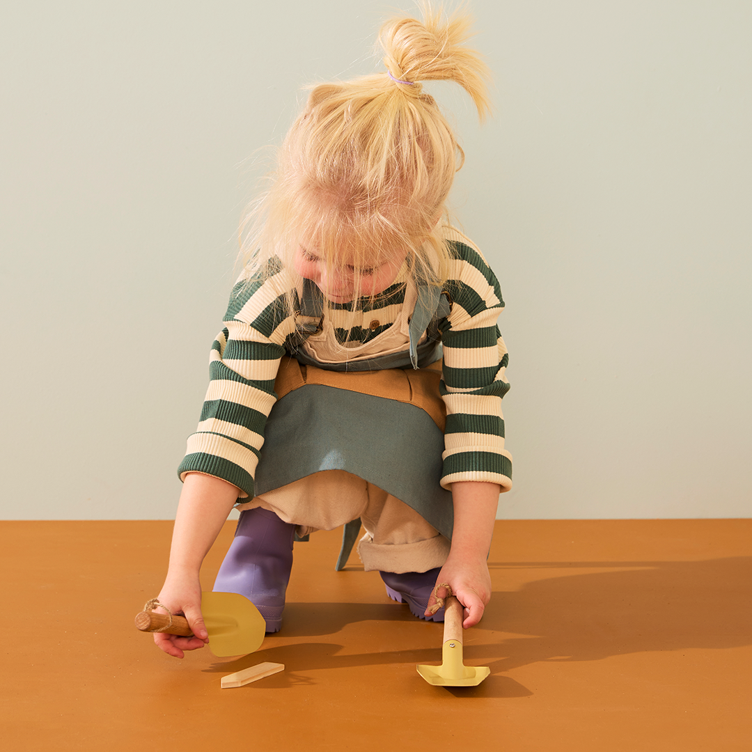 Een meisje met blond haar knielt en speelt met een geel schepje en harkje van de Kids Concept tuingereedschapsset.