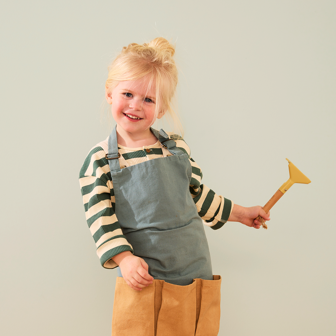 Een lachend meisje met een blauw schort houdt een geel harkje van de Kids Concept tuingereedschapsset vast.