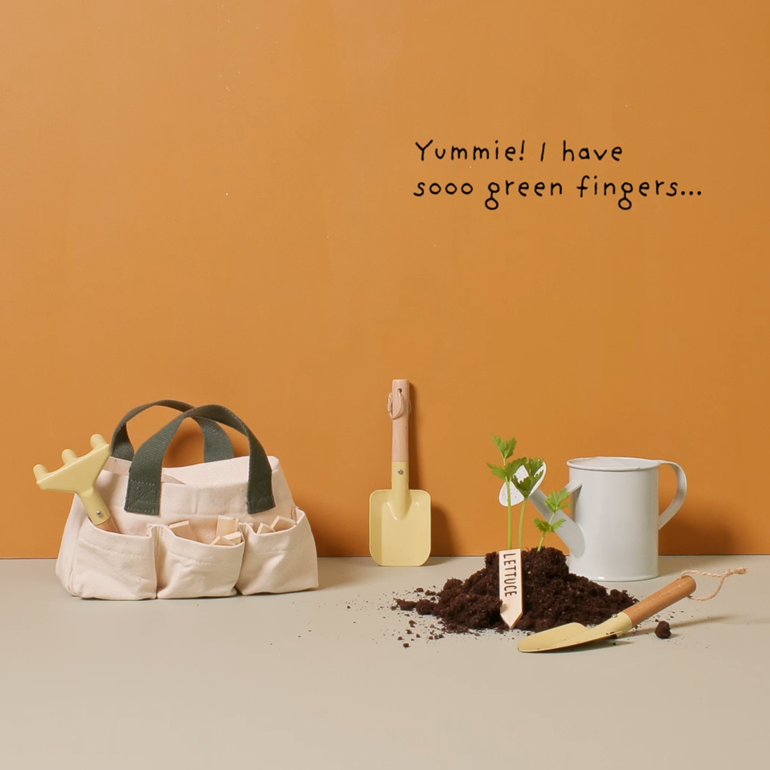De Kids Concept tuingereedschapsset met tas, schepje, gieter en een plantje in aarde op een tafel.