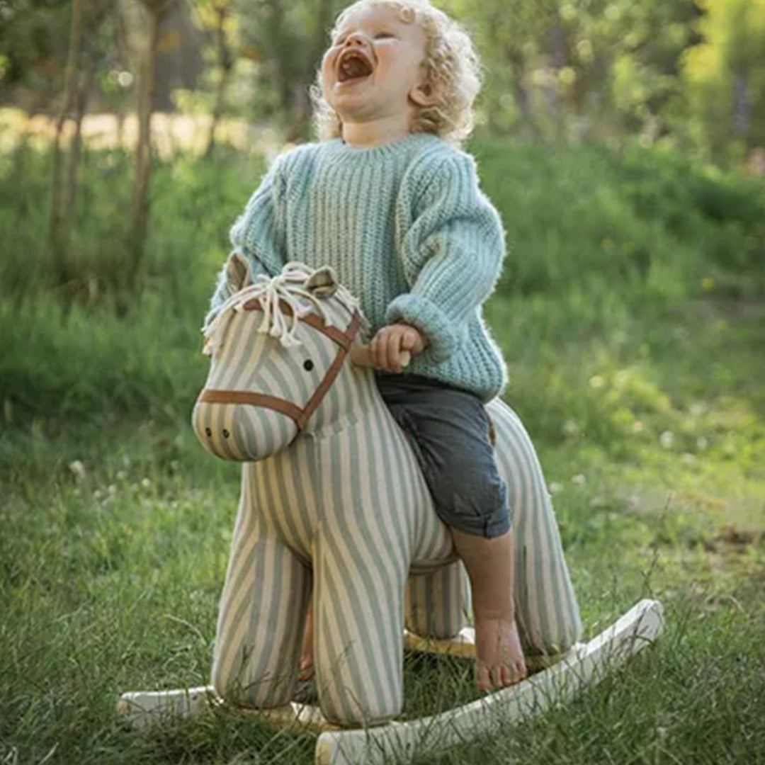 Een lachend kind zit op het Kids Concept gestreepte hobbelpaard in het gras.