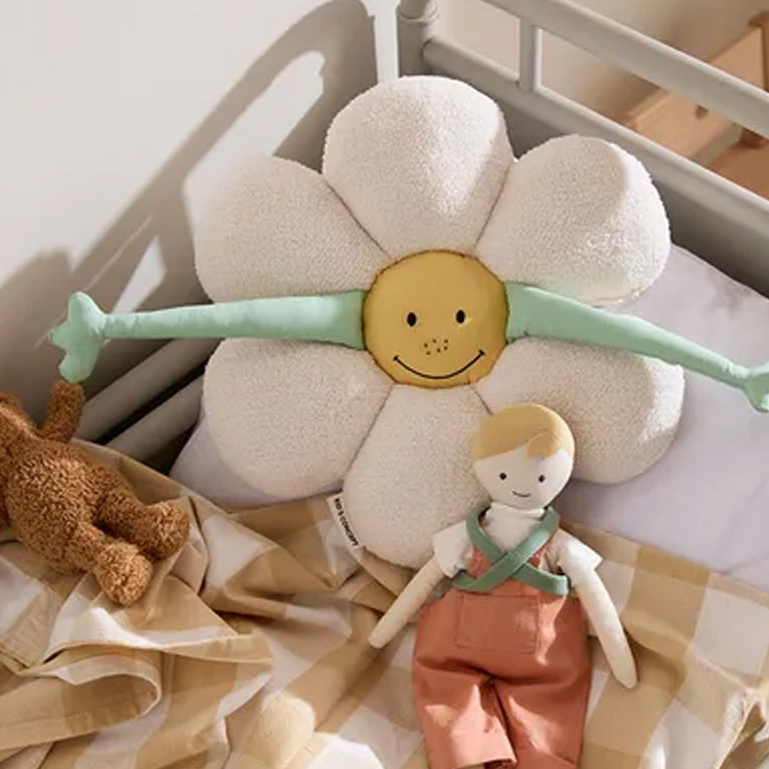 Een kids concept bloemkussen met een geel vrolijk gezicht ligt op een bed naast een teddybeer en een pop.