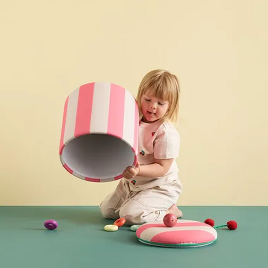 Een kind knielt en houdt de kids concept opbergpoef met roze en witte strepen vast, met de deksel en speelgoed op de grond.