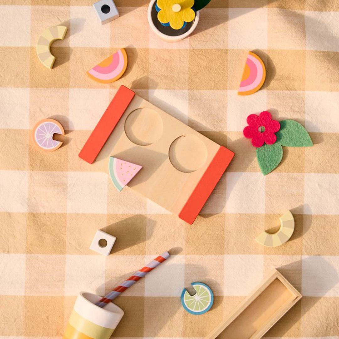 Kids Concept houten drankenset onderdelen, waaronder bekers, fruit en een dienblad, verspreid over een geel geruite deken.