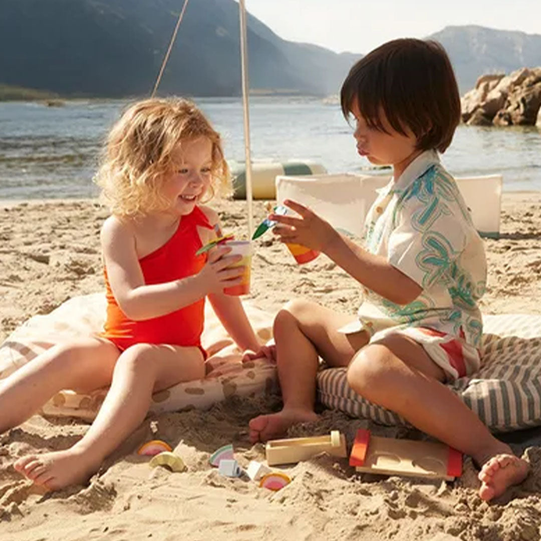 Twee kinderen spelen met de Kids Concept houten drankenset op een zandstrand met bergen en water op de achtergrond.