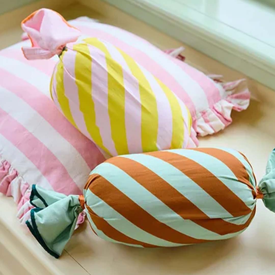 Twee Kids Concept snoepkussens, roze-geel en bruin-mint, liggen op een bed met een roze gestreept kussen.