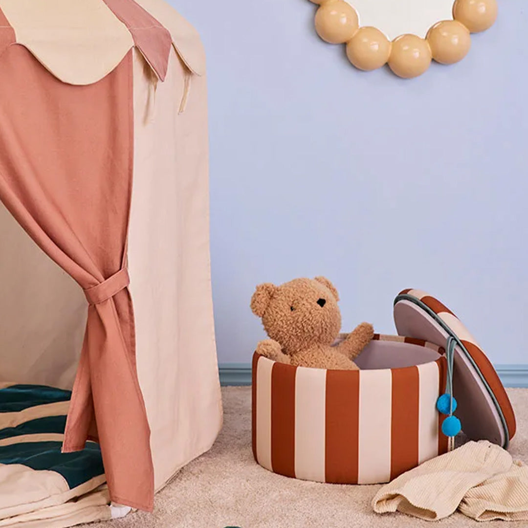 Een teddybeer zit in de open kids concept gestreepte opbergpoef naast een speeltent.