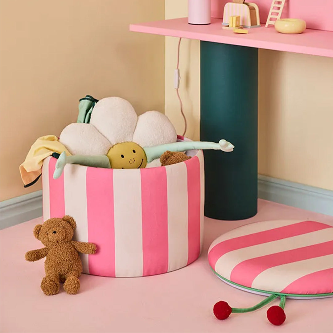 kids concept opbergpoef met roze en witte strepen, gevuld met speelgoed, naast een teddybeer in een kinderkamer.