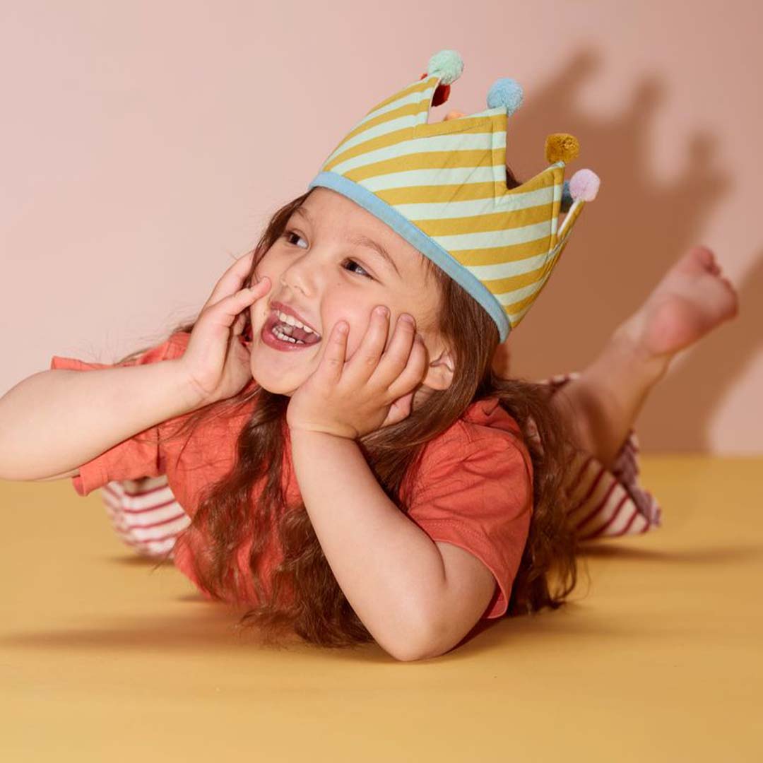 Een lachend kind ligt op een gele ondergrond en draagt de Kids Concept verjaardagskroon met gestreepte binnenkant en pompons.