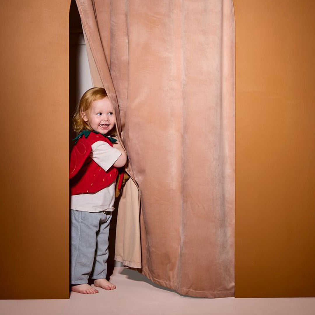 Een lachend kind draagt de Kids Concept aardbeienfee verkleedkleding en kijkt achter een gordijn vandaan.