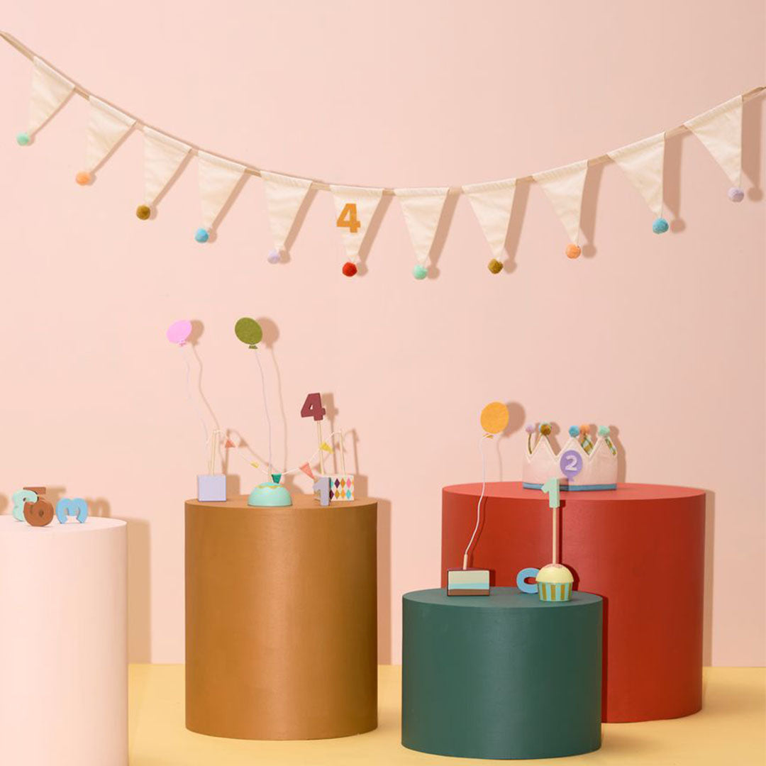Kids Concept stoffen vlaggenslinger met beige vlaggetjes en kleurrijke pompons hangt boven diverse verjaardagsdecoraties op roze en bruine sokkels.