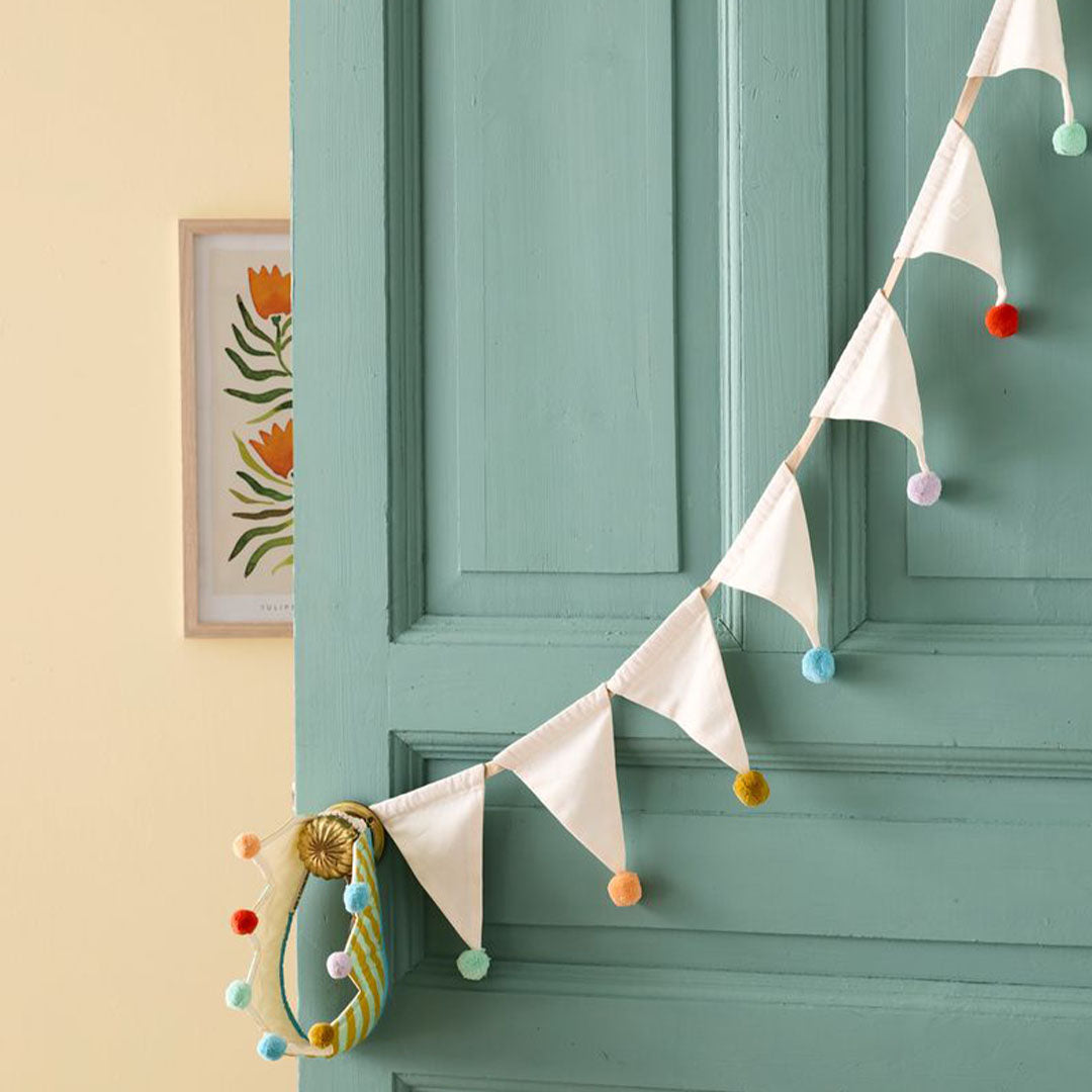 Kids Concept stoffen vlaggenslinger met beige vlaggetjes en kleurrijke pompons hangend aan een teal deur.
