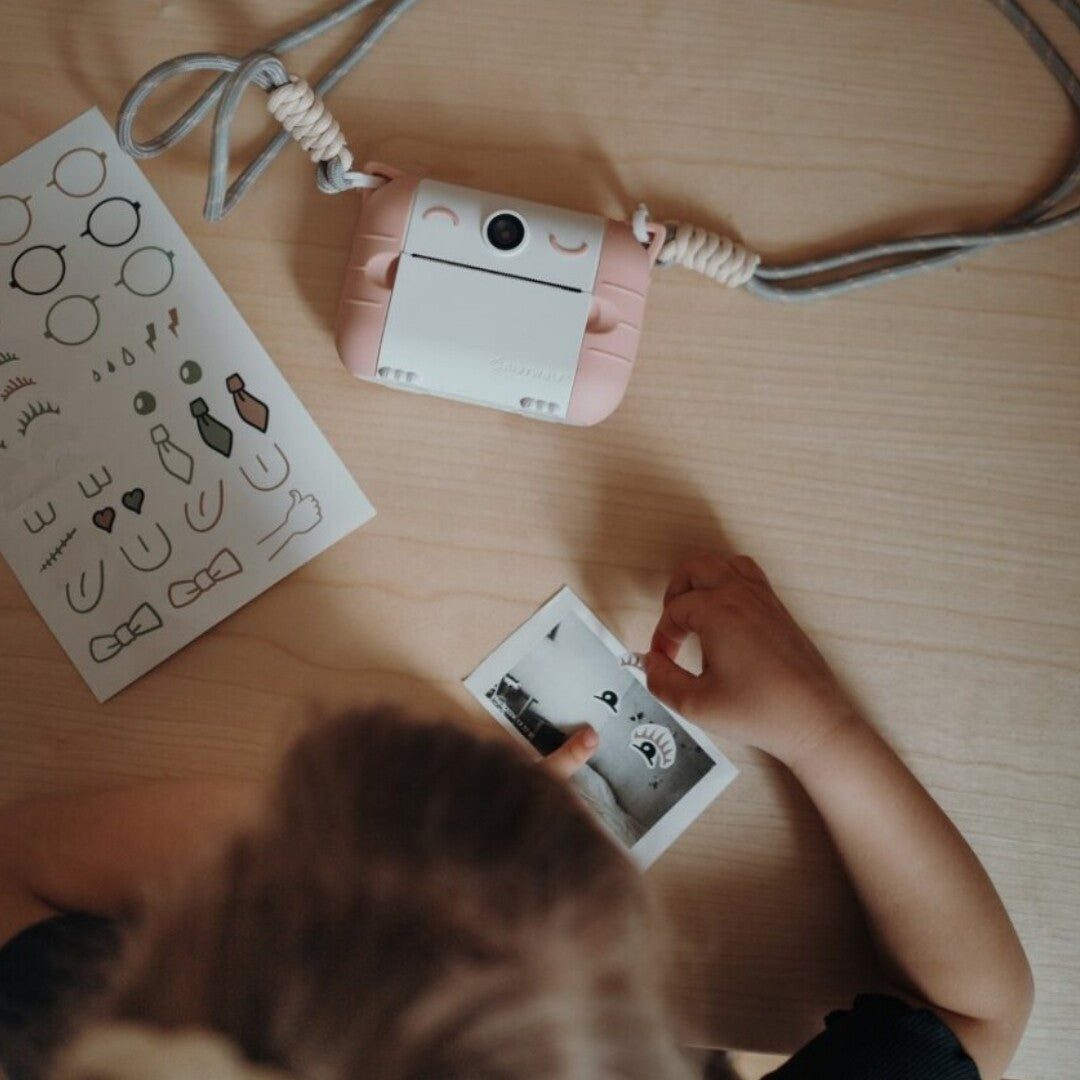 Een kind decoreert een kleine foto met stickers, naast een roze-witte kidywolf kindercamera en een stickervel op een houten tafel.