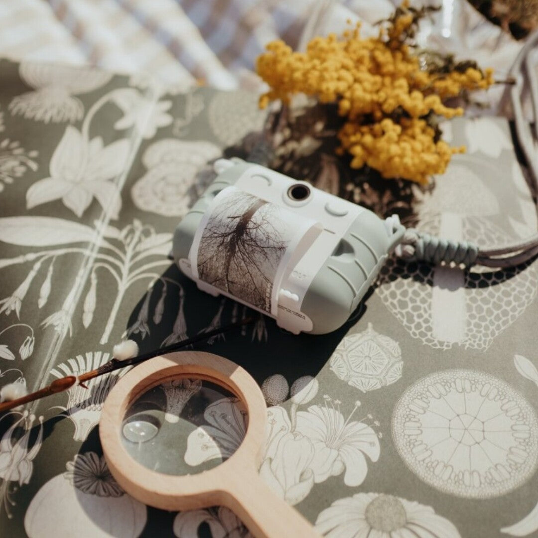 Een lichtblauwe kidywolf kindercamera met een geprinte boomfoto ligt naast een houten vergrootglas en gele bloemen.