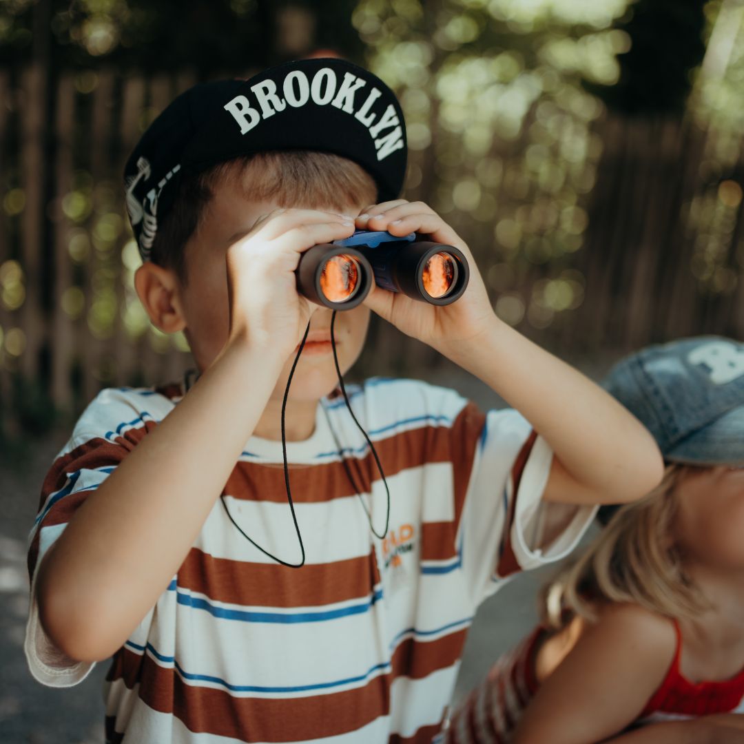 Een kind met een 'Brooklyn' pet kijkt door een blauwe Kidywolf verrekijker.