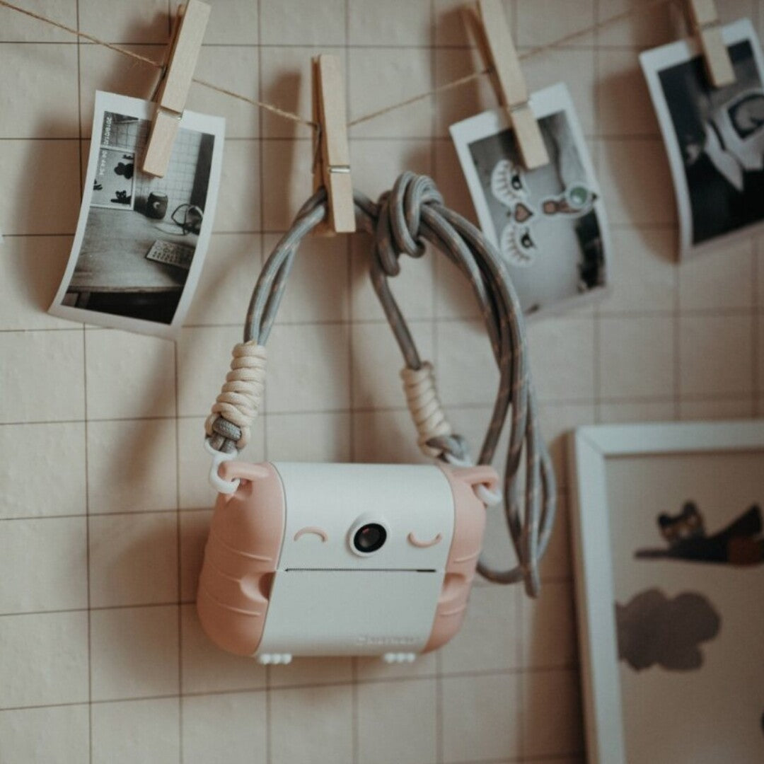 Een roze kidyprint kindercamera hangt aan een touw aan de muur, met geprinte foto's erboven.