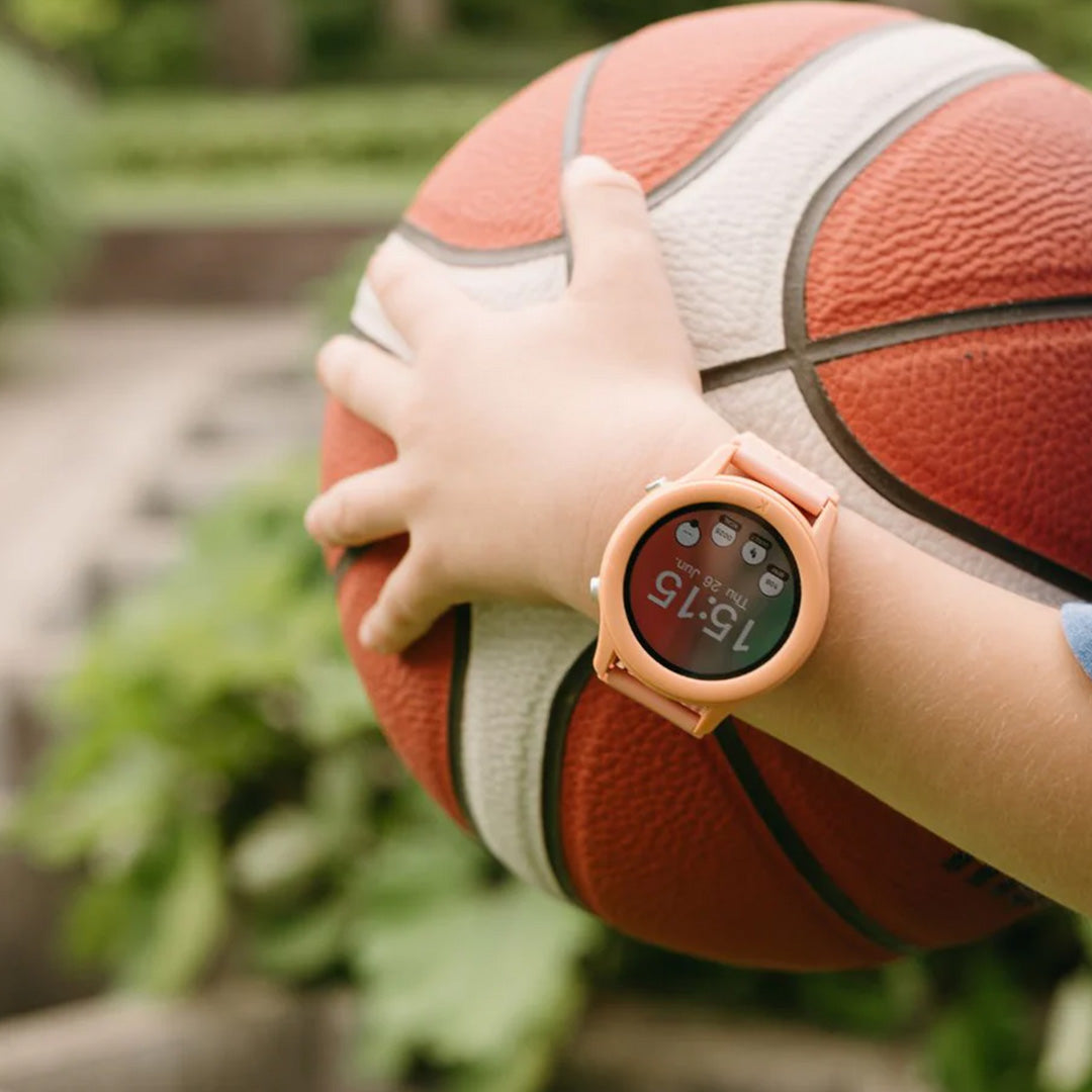 Een kind draagt het Kidywolf K-Watch interactief horloge in koraal kleur terwijl het een basketbal vasthoudt.