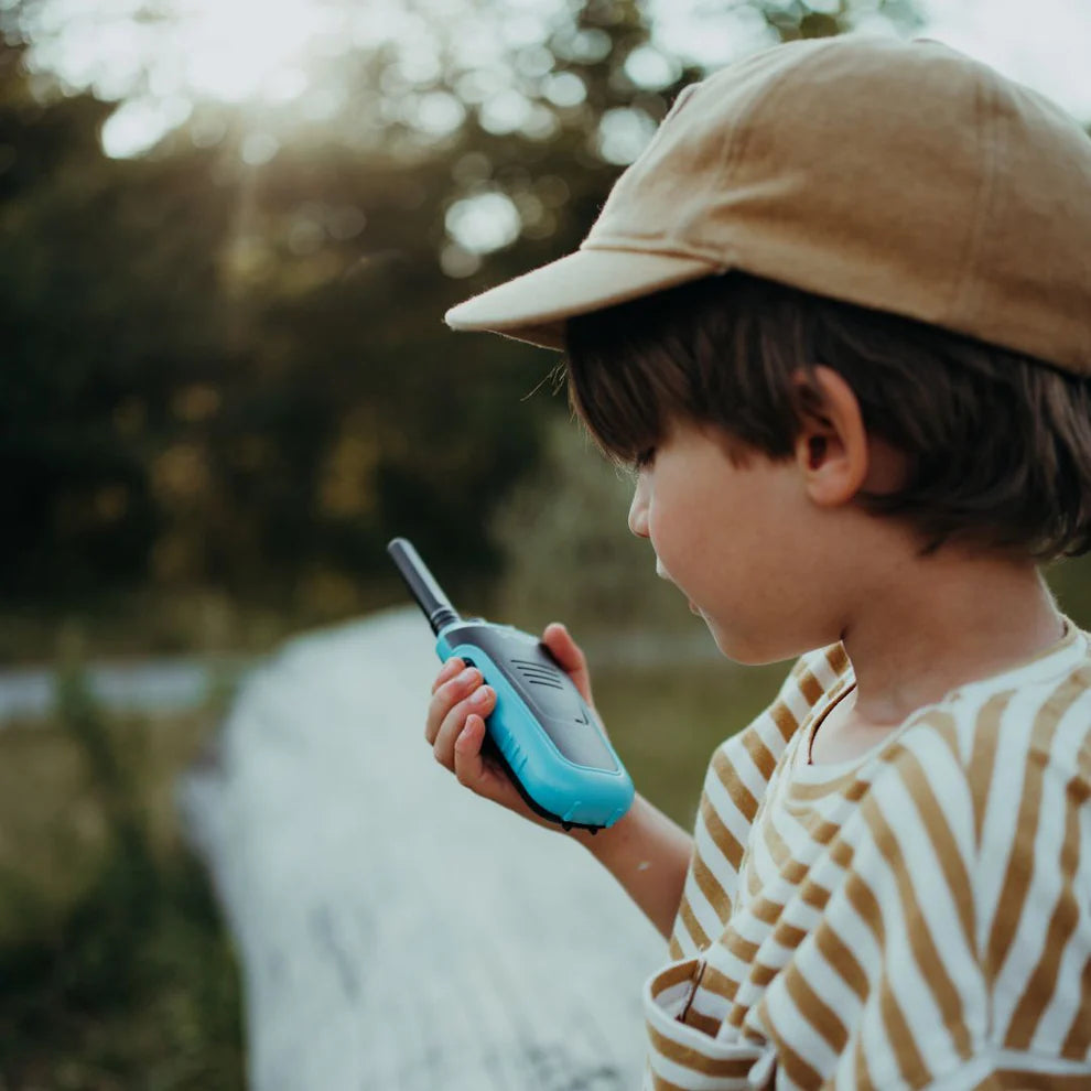 Een kind met een pet kijkt naar de groene kidywolf Kidytalk walkie talkie die het vasthoudt, buiten in de zon.