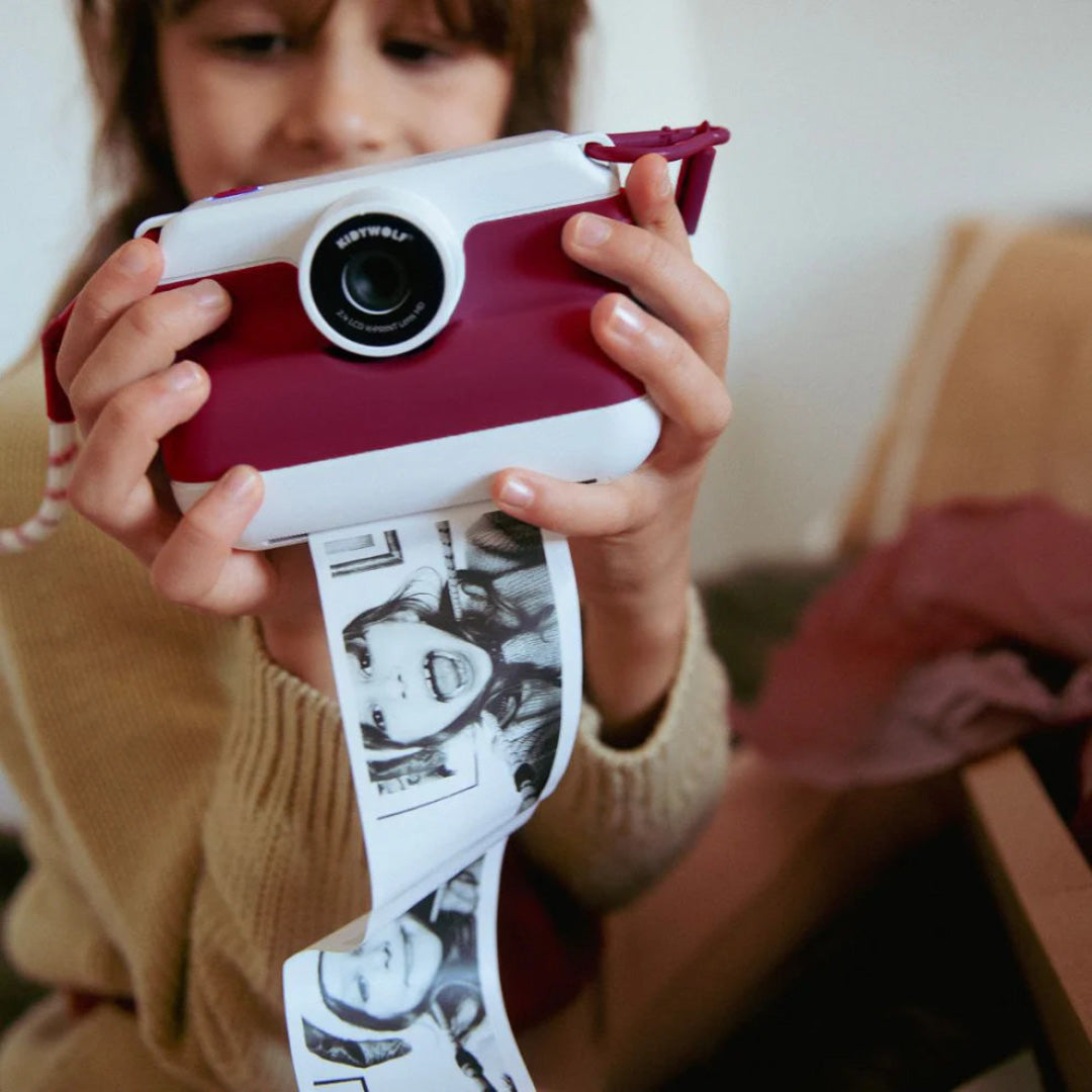 Een kind houdt de rode Kidywolf kindercamera vast waar een lange strook zwart-wit foto's uitkomt.