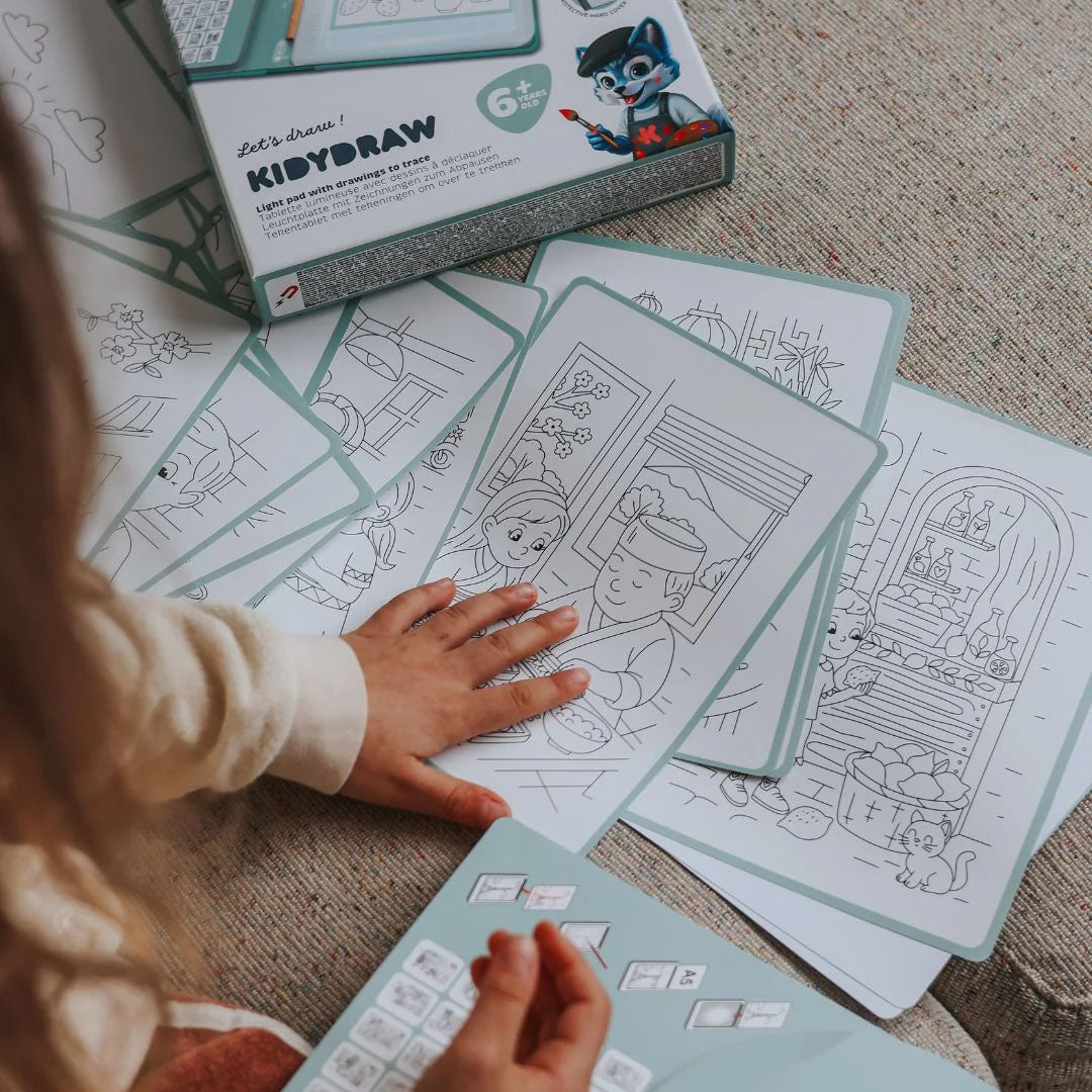Een kinderhand kiest uit verschillende zwart-wit tekensjablonen met figuren en eten, naast het Kidywolf KidyDraw tekenbord.