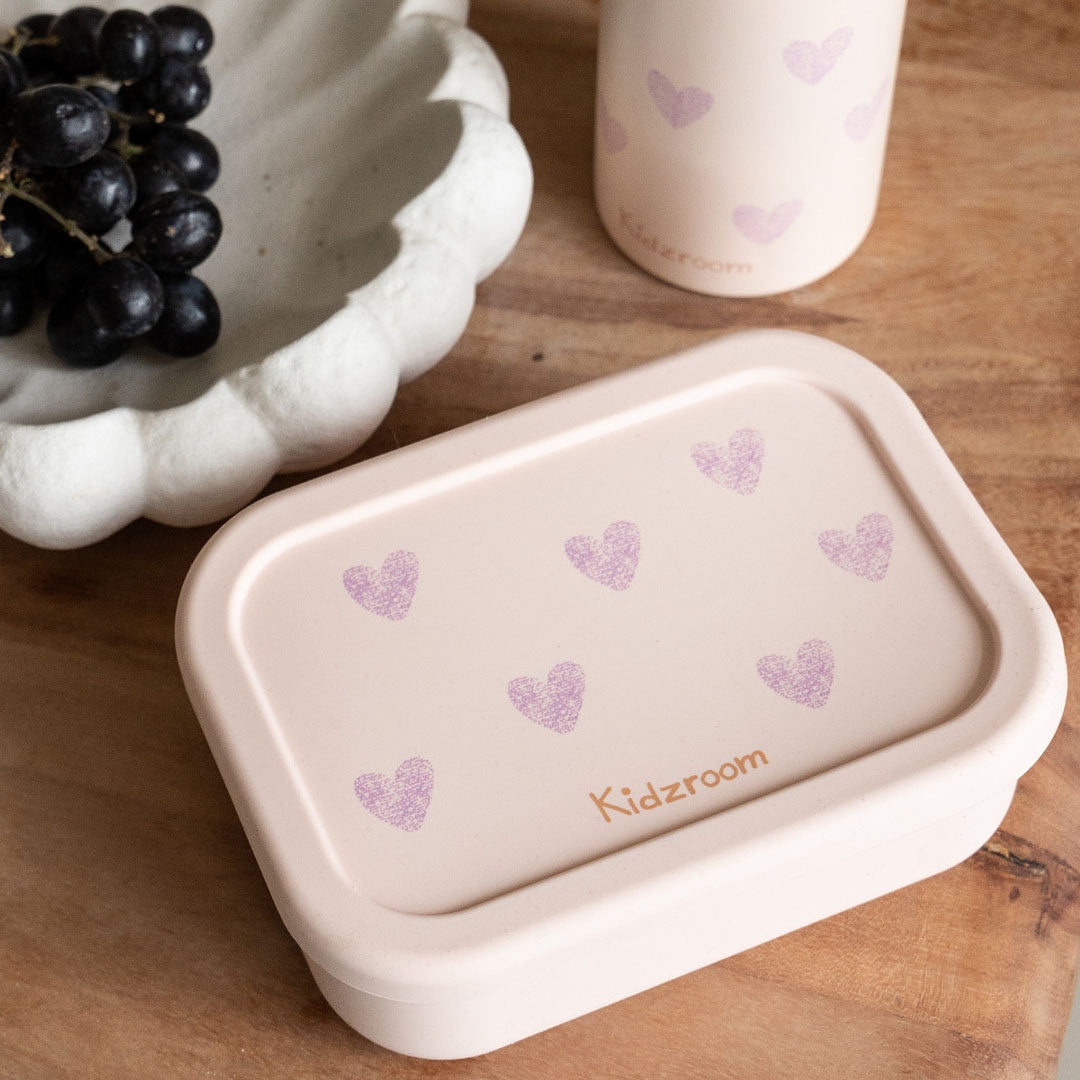 Kidzroom roze broodtrommel met hartenpatroon op een houten ondergrond, met druiven en een drinkfles op de achtergrond.