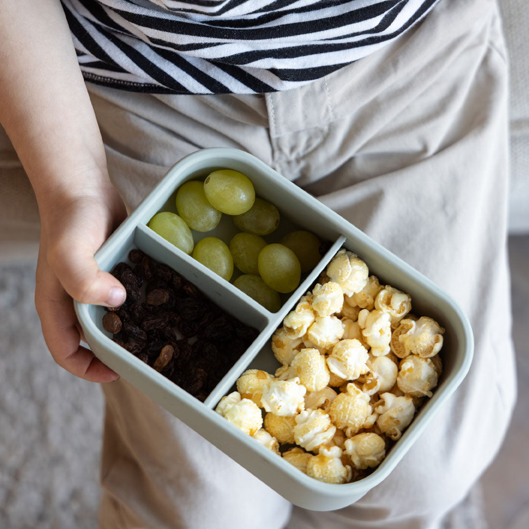 Een kind houdt een open broodtrommel vast gevuld met druiven, rozijnen en popcorn.