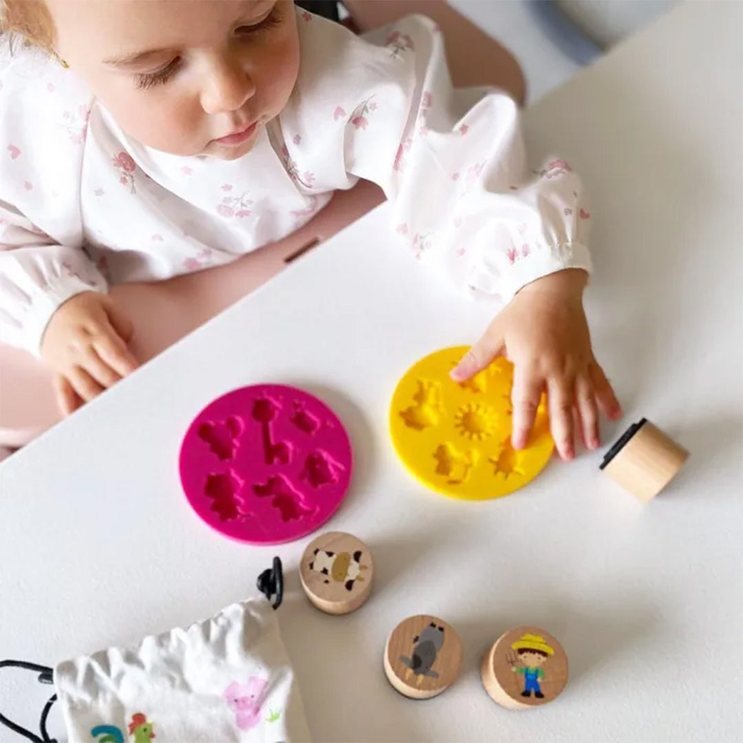 Een peuter speelt met roze en gele siliconen vormen met dierfiguren en houten stempels op een witte tafel.