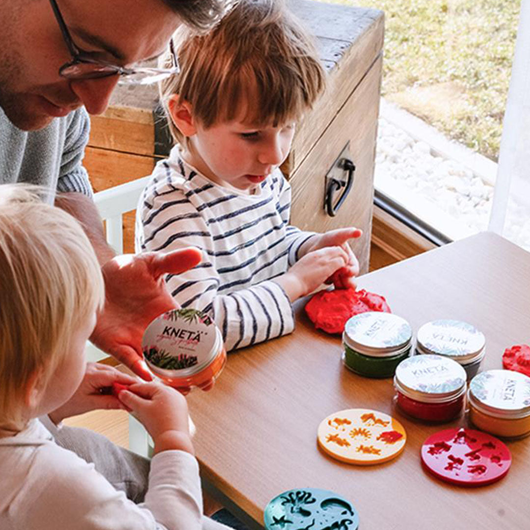 Een vader en twee kinderen spelen aan tafel met Knetä speelklei en verschillende vormpjes.