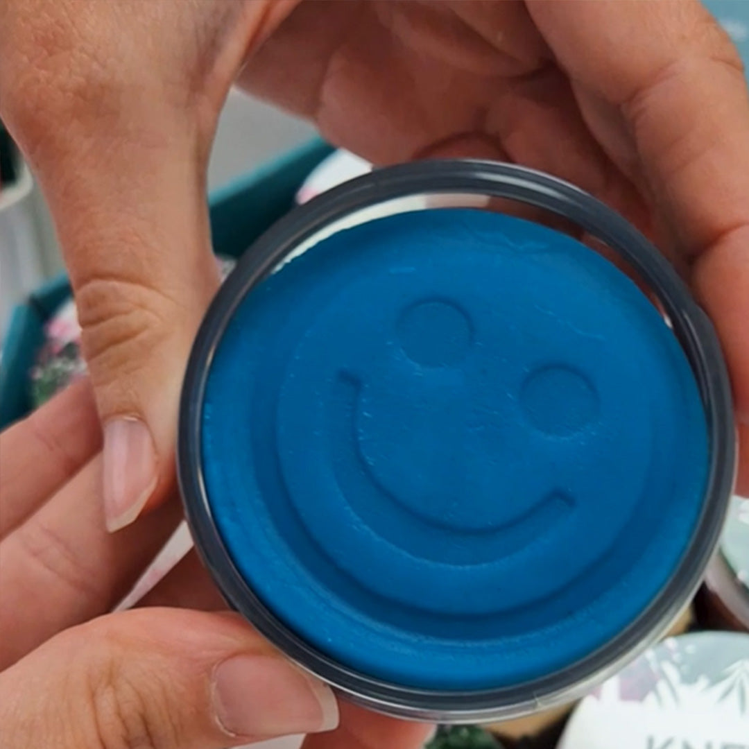 Detail van handen die een blauw Knetä speelklei deksel met een smiley vasthouden.