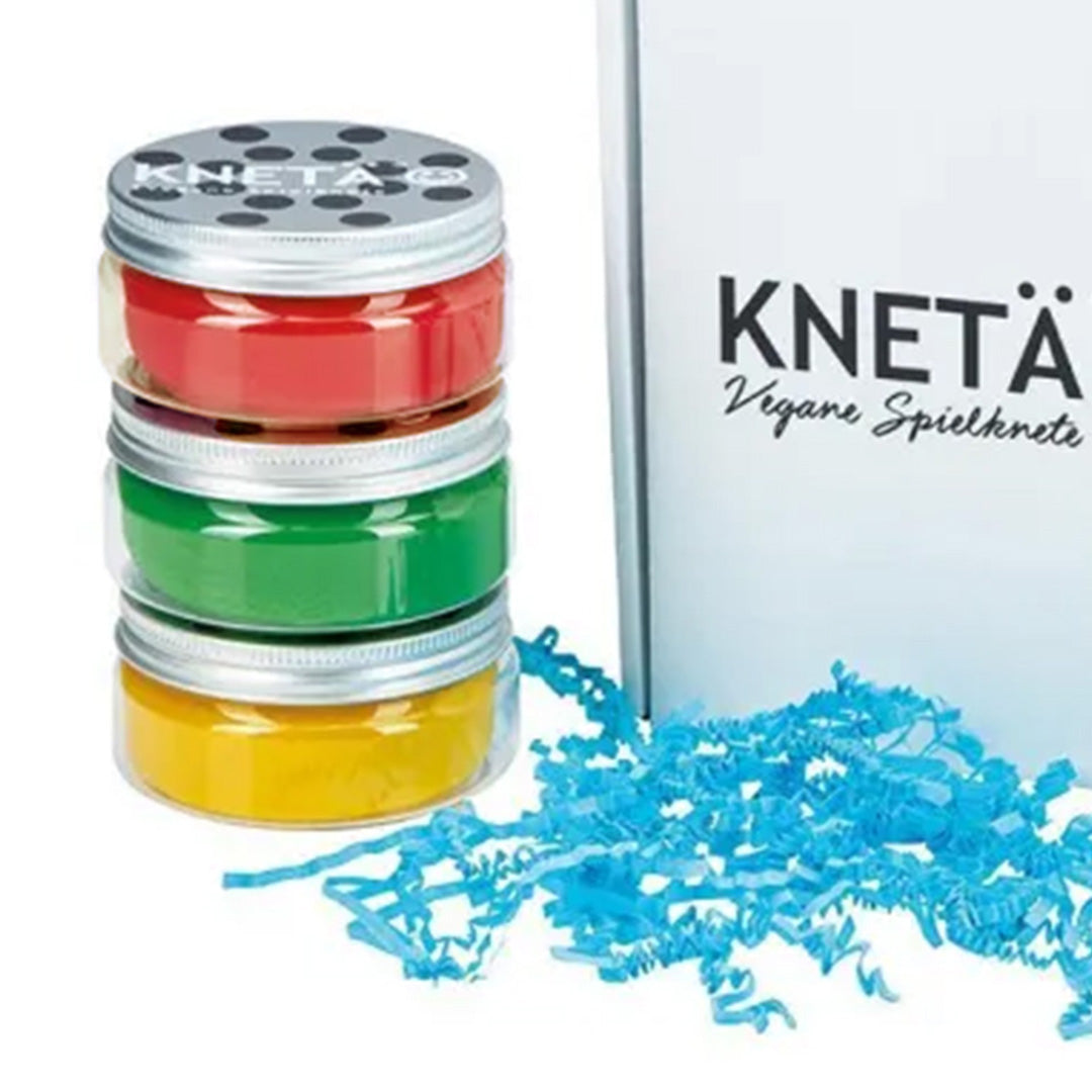 Drie gestapelde potjes Knetä speelklei in rood, groen en geel naast een witte doos en blauw snipperpapier.