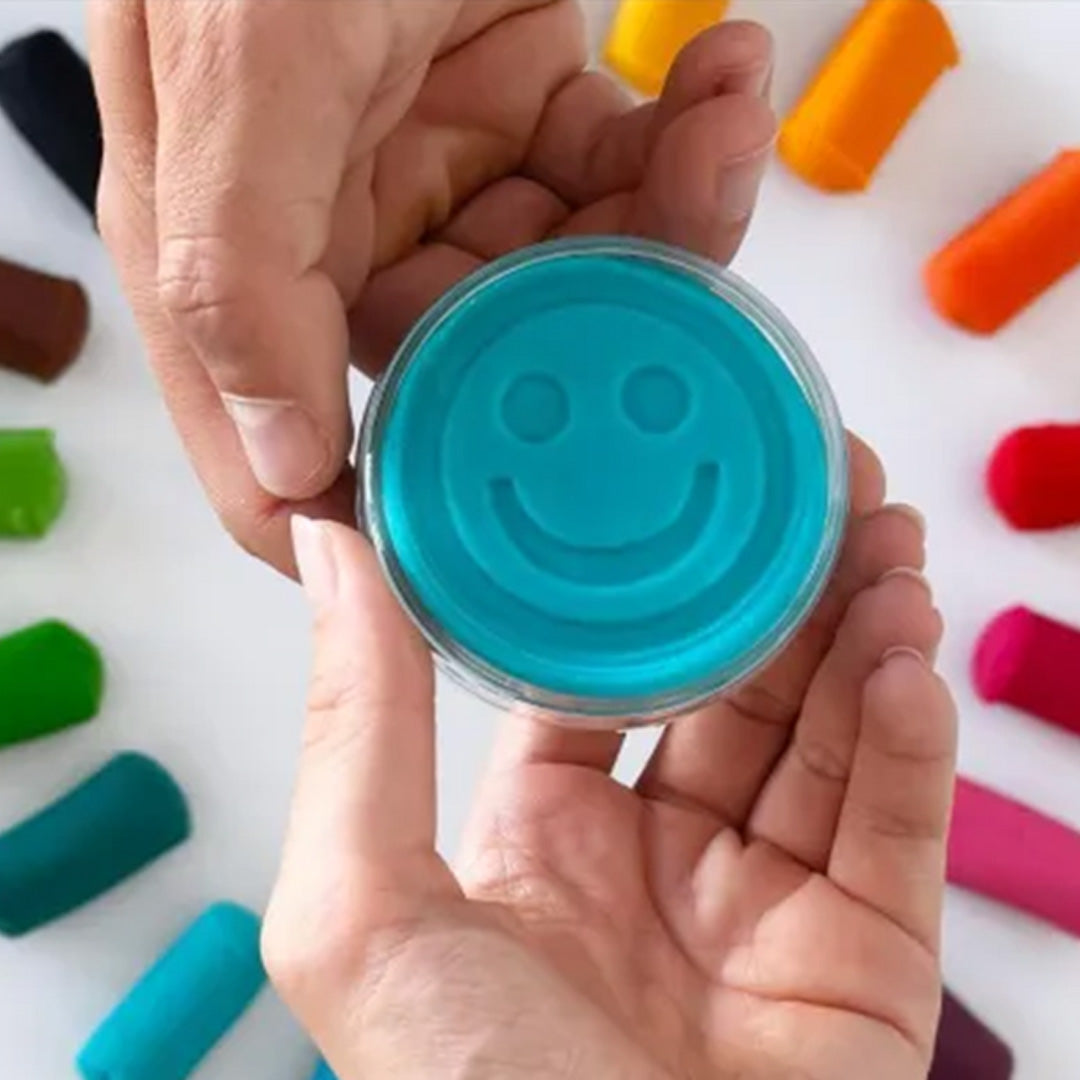 Twee handen houden een rond potje blauwe Knetä speelklei vast met een smiley-afdruk op de bovenkant.