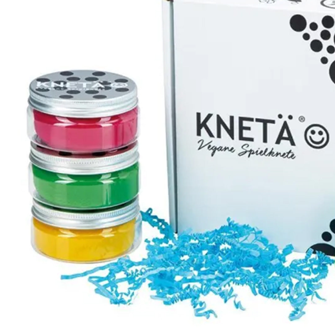 Drie gestapelde potjes Knetä speelklei in roze, groen en geel, naast een witte doos en blauw snipperpapier.