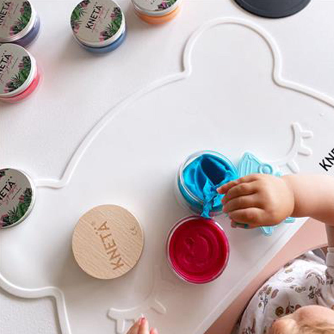 Een kinderhand speelt met blauwe en roze Knetä speelklei op een wit speelmatje in de vorm van een beer.