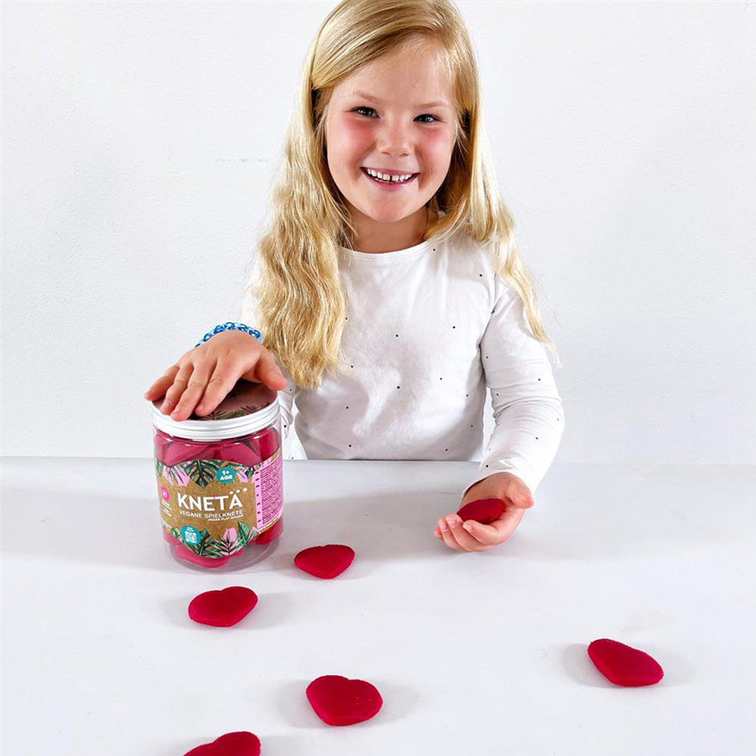 Een lachend meisje speelt met Knetä speelklei in de vorm van roze hartjes op een witte tafel.